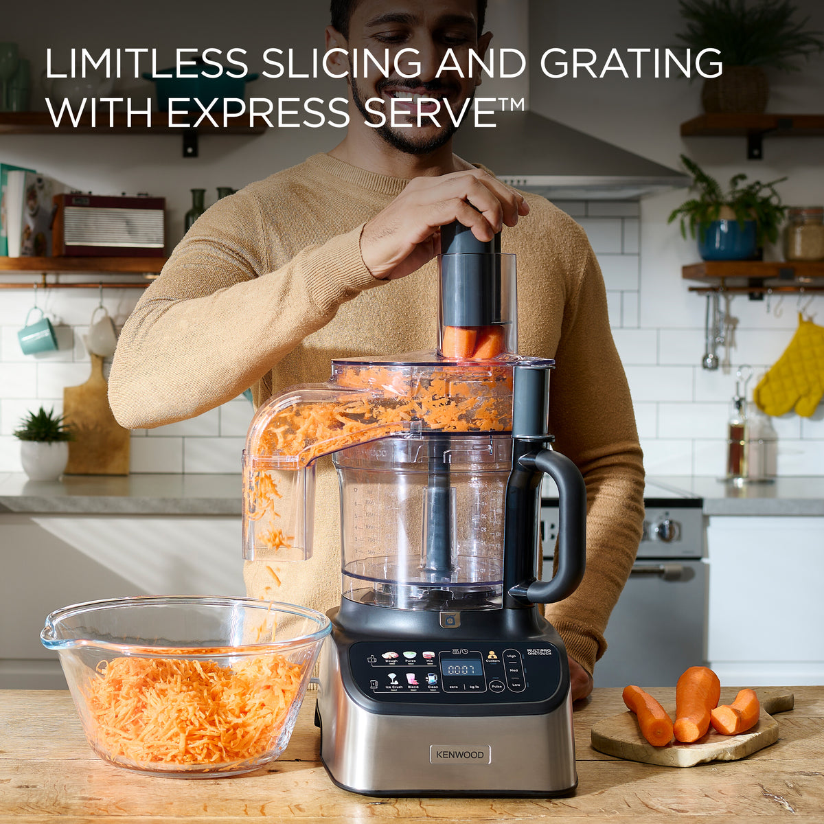 Kenwood 7-in-1 Multipro Express food processor grating carrots into a glass bowl on a kitchen counter.