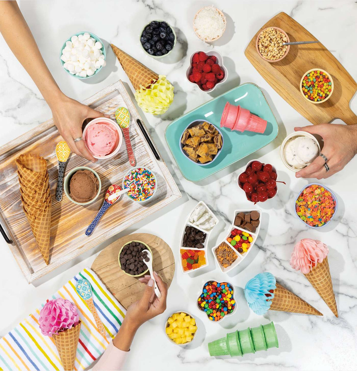 Hands holding cups of ice cream surrounded by cones, toppings, and colorful spoons on a marble surface