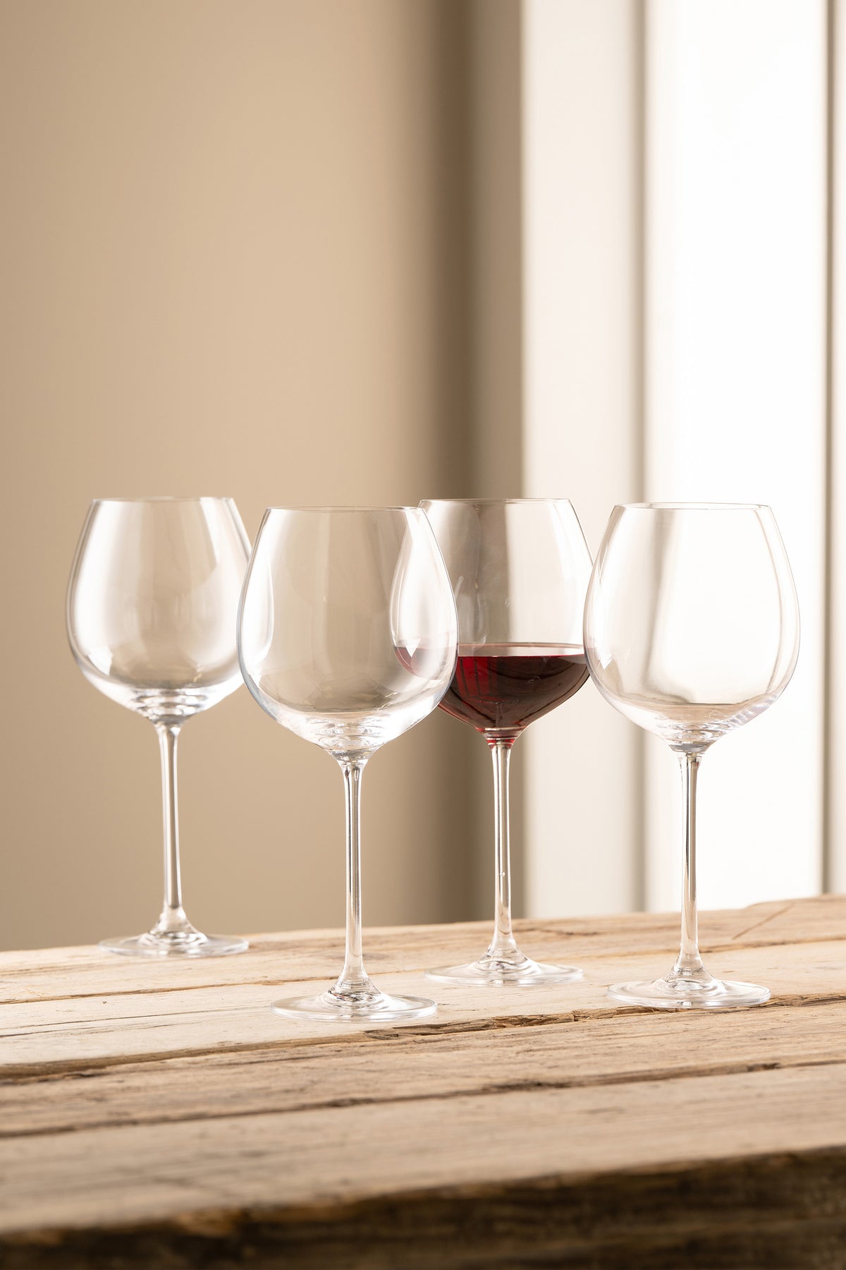 Four clear Solus Goblet wine glasses on a wooden surface, one partially filled with red wine