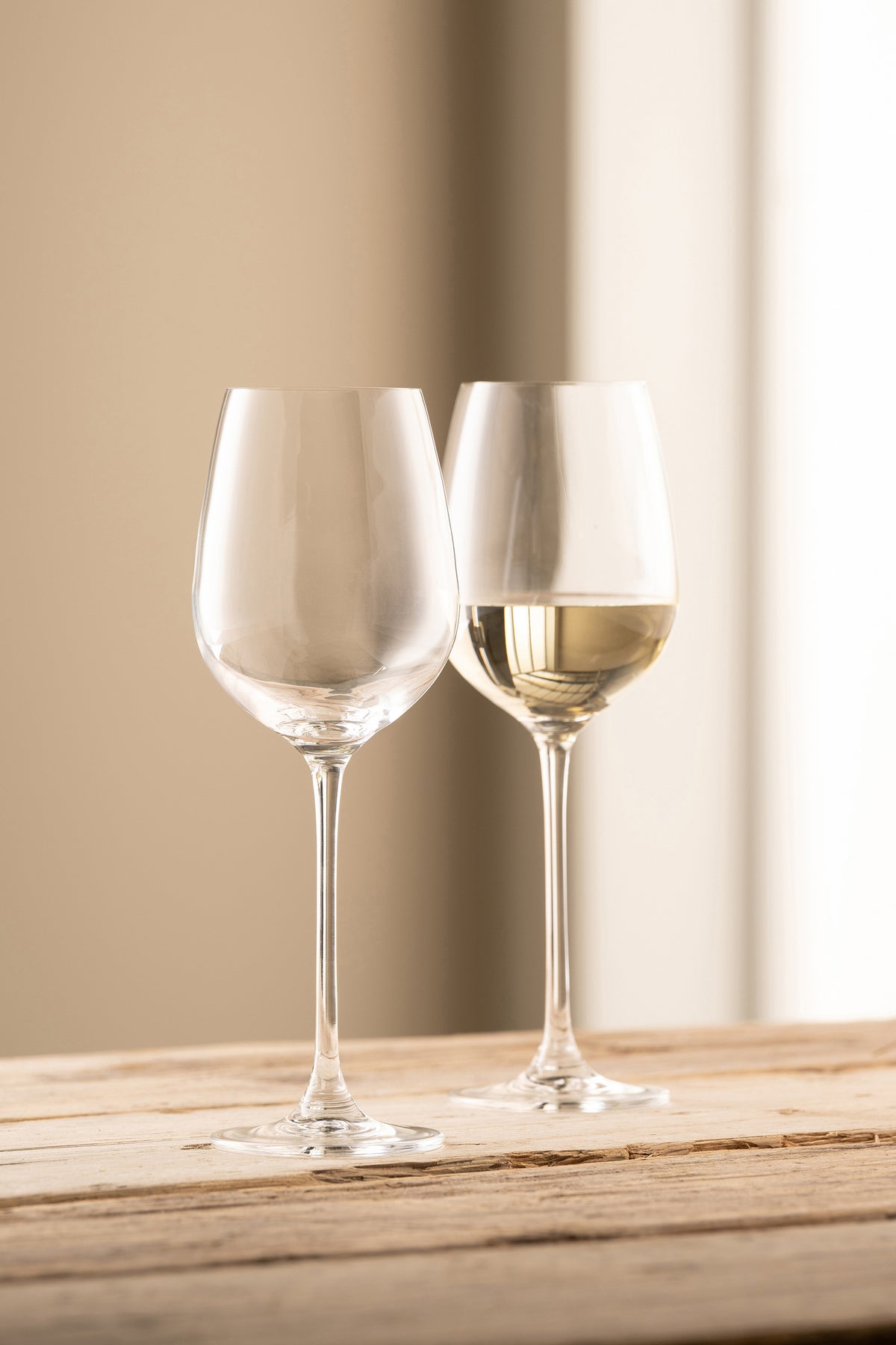 Two clear Solus Wine glasses on a wooden surface, one filled with white wine, against a beige background.
