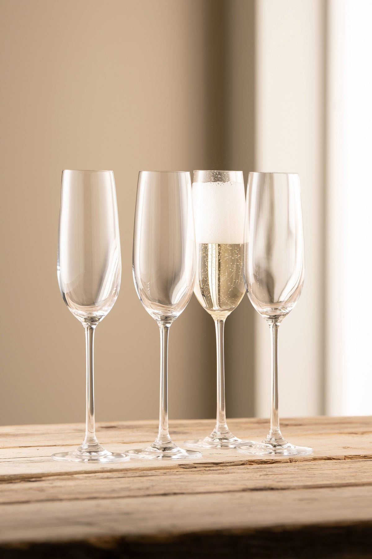 Four clear Solus Flute glasses on a wooden surface, one filled with sparkling champagne and foam.