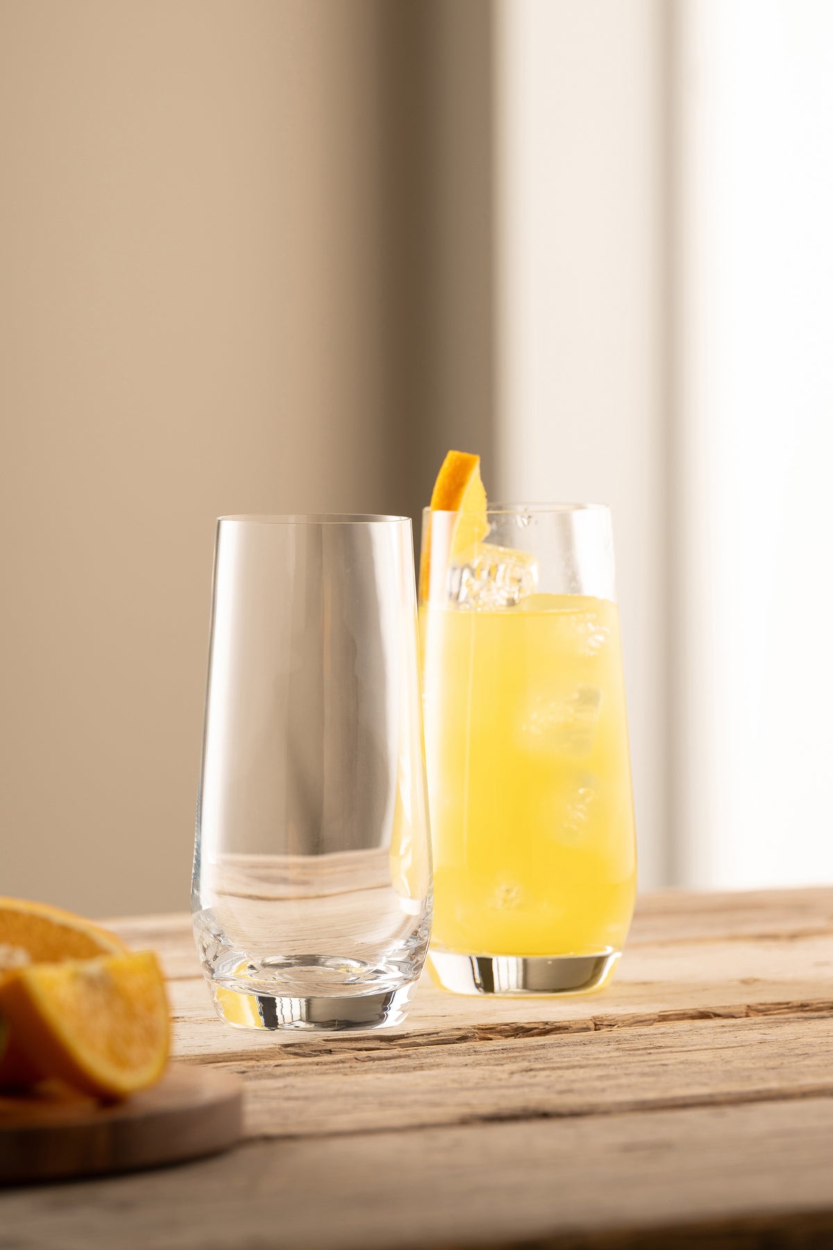 Two clear Solus Hi Ball glasses on a wooden surface, one filled with orange drink and ice, garnished with orange slice.