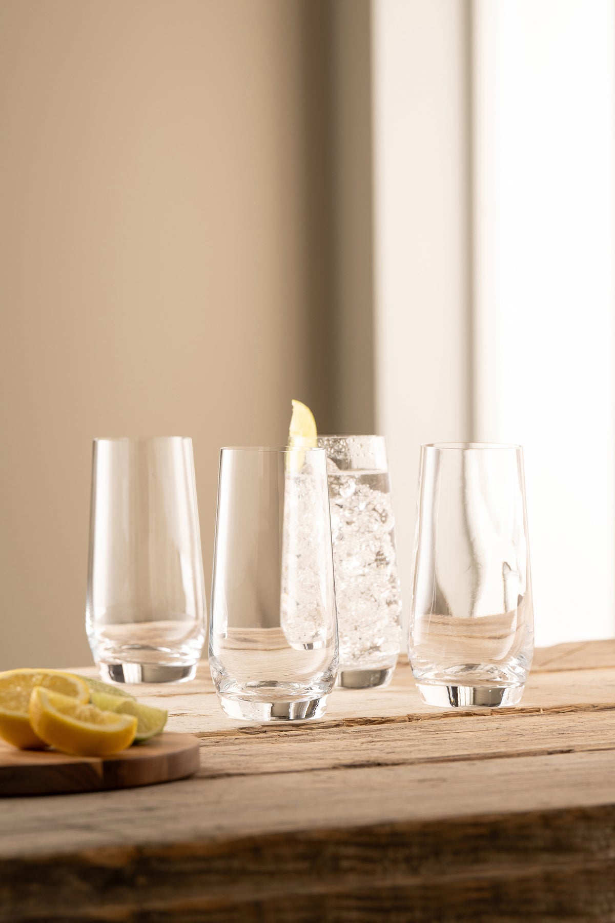 Four clear Solus Hi Ball glasses on a wooden surface, one filled with ice and a lemon wedge, lemon slices nearby.