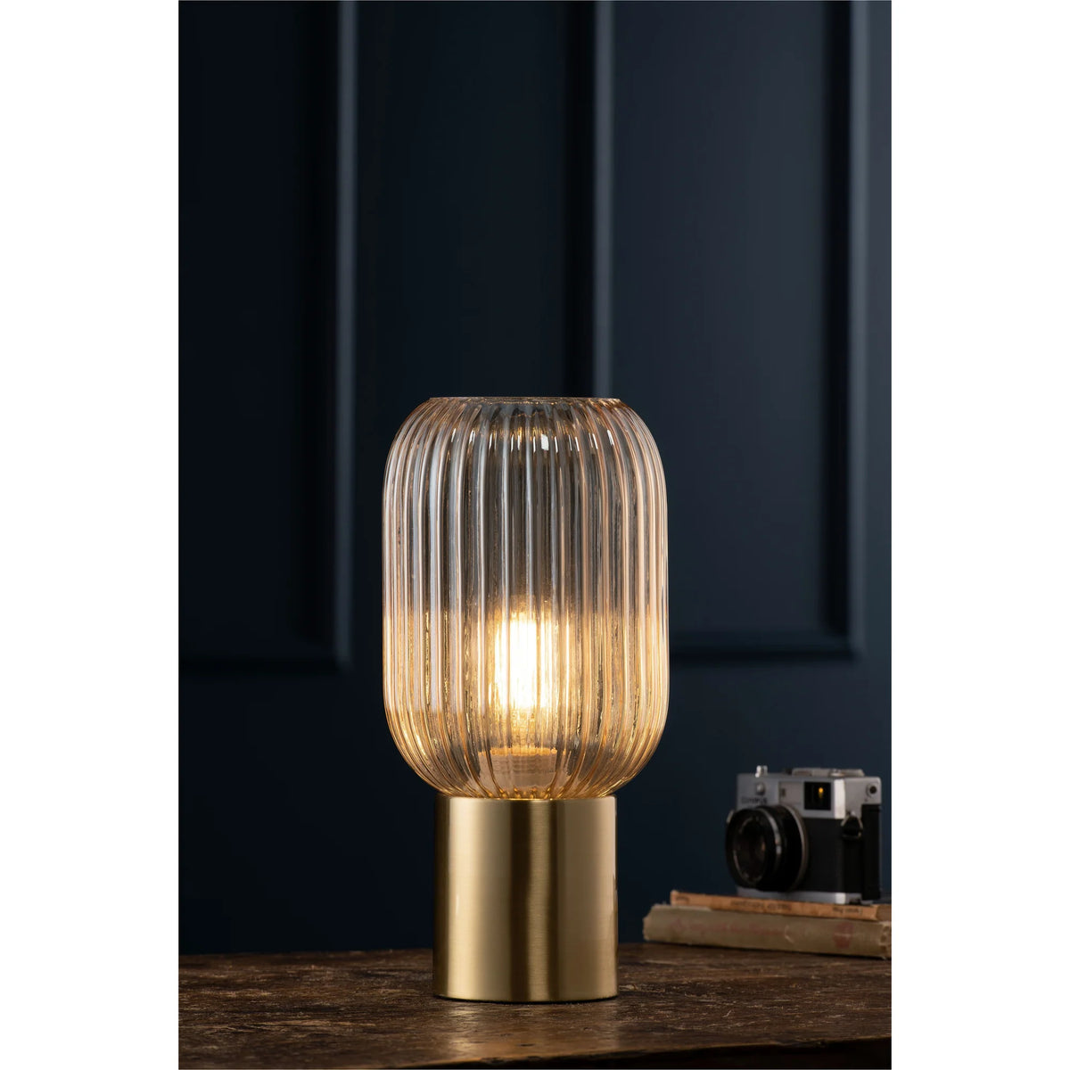 Fluted amber glass table lamp with brass base lit on a wooden surface, dark wall background.