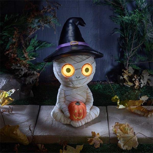 Halloween-themed mummy decoration with glowing eyes and a witch hat, sitting on a stone surface with autumn leaves.