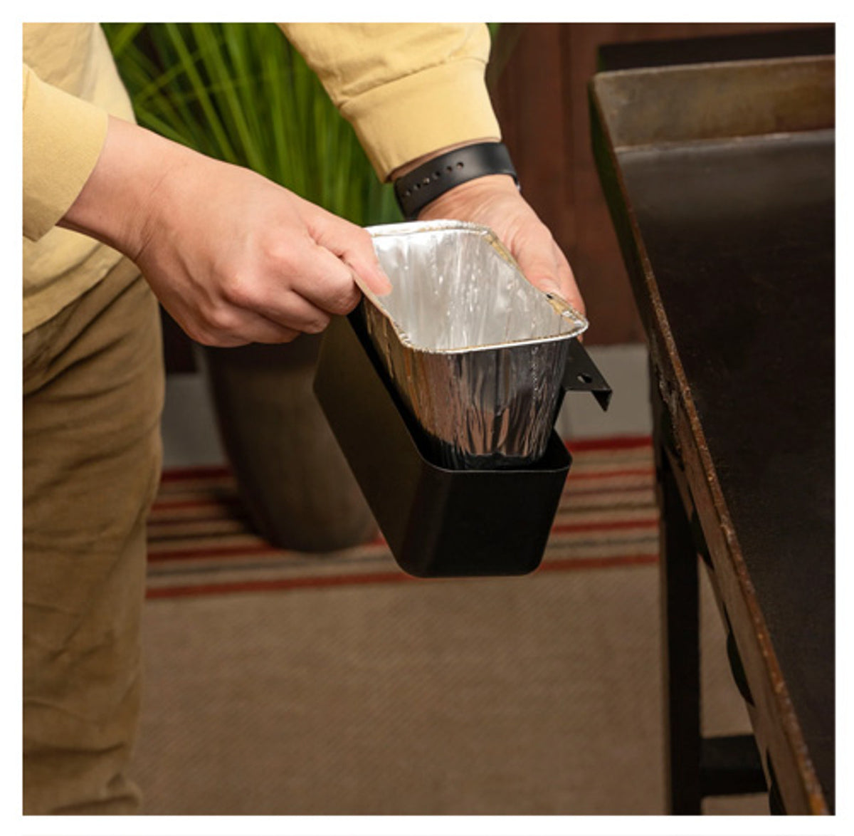 Hands placing a foil liner into a black Blackstone grease cup attached to a griddle side shelf