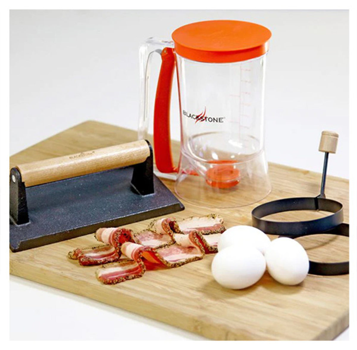 Blackstone Breakfast Kit with clear batter dispenser, bacon press, egg rings, and three eggs on wooden board