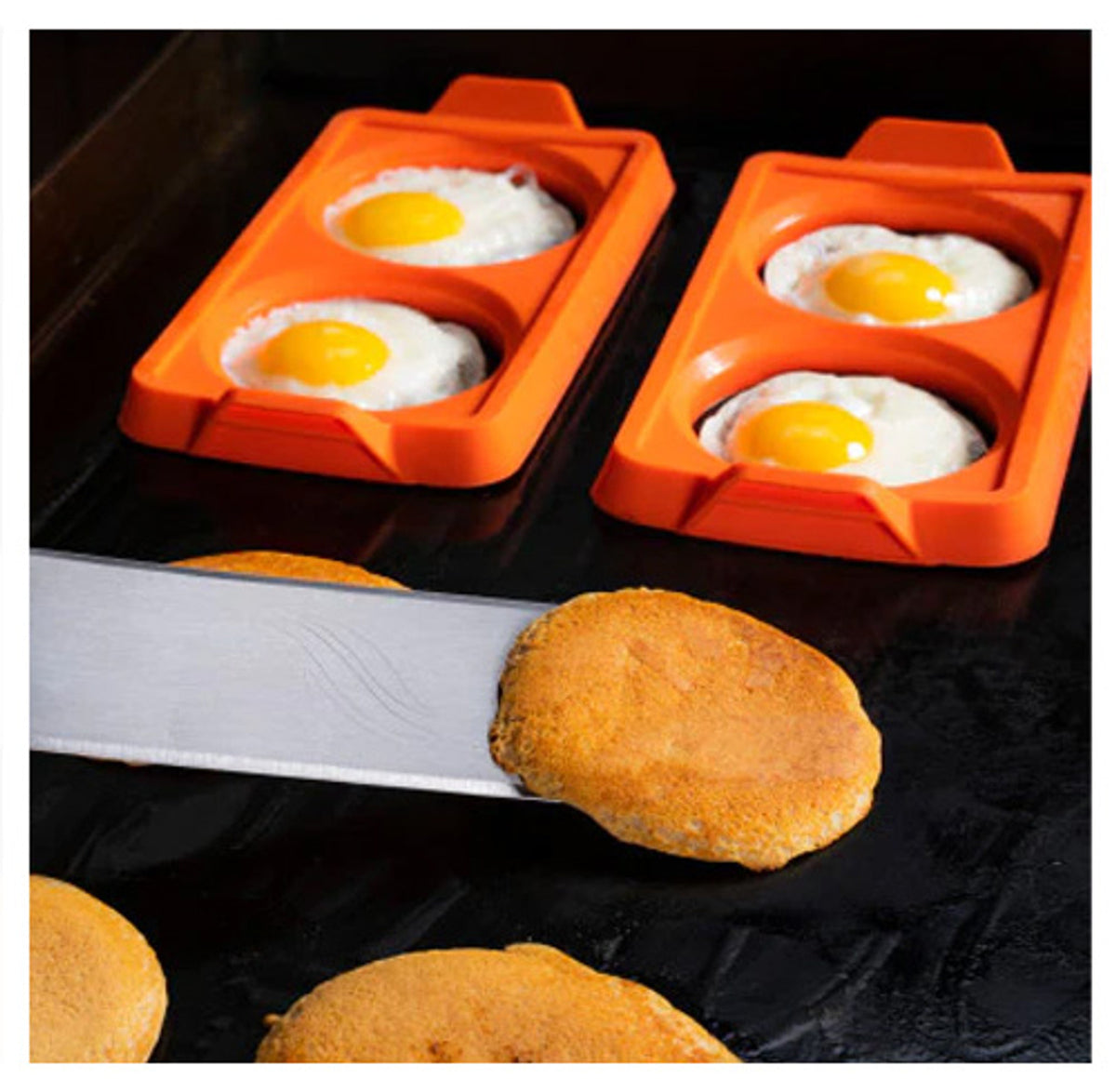 Two orange Blackstone 2 Section Egg Ring Trays cooking eggs on a griddle with pancakes nearby.