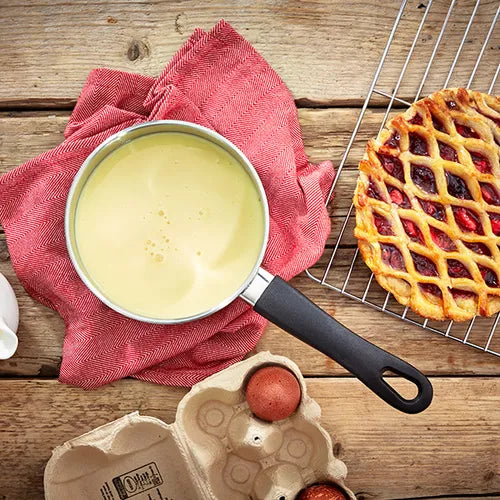 Judge Vista 14cm milk pan with creamy sauce inside, resting on a red cloth on a wooden surface next to eggs and a lattice pie