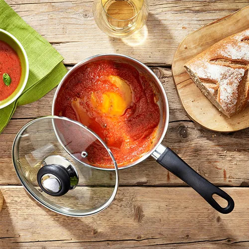 Judge Vista 18cm saucepan with tomato sauce and melted cheese, glass lid beside it on wooden table with bread and drink