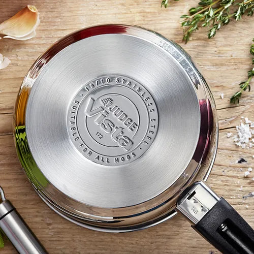 Bottom view of Judge Vista 20cm saucepan showing stainless steel base and handle on wooden surface with herbs and garlic.