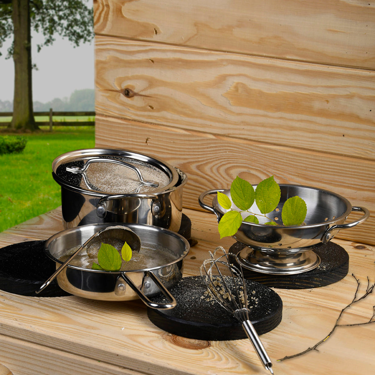 Mud Kitchen Toolset 6 Piece with metal pots, colander, whisk, and ladle on wooden surface with leaves and sand.