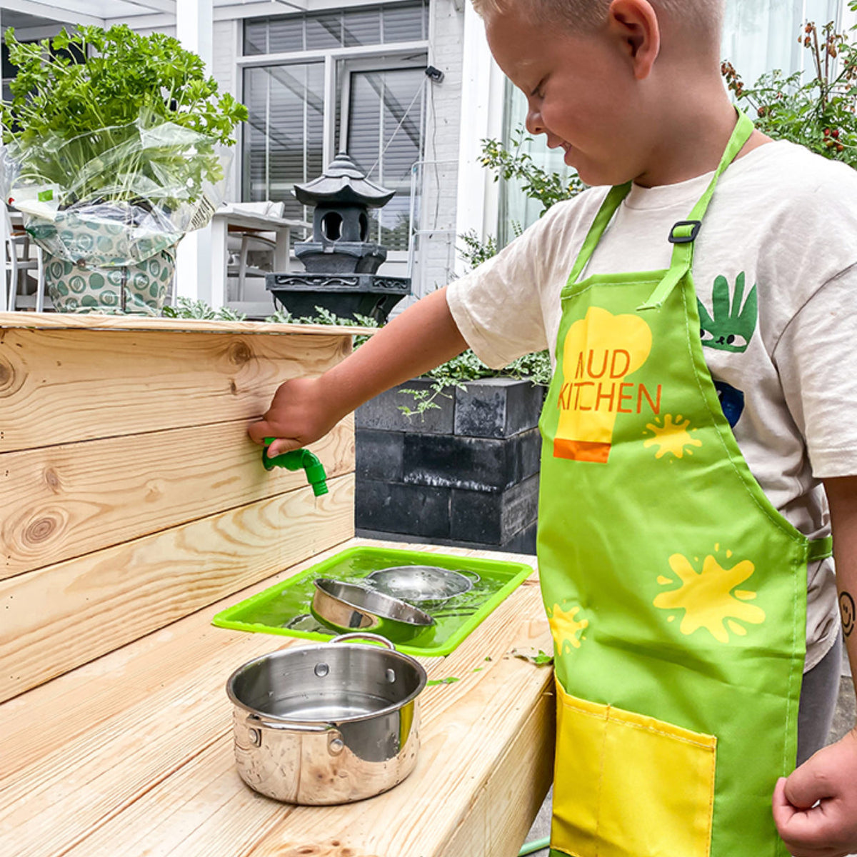 Child wearing a green Mud Kitchen apron playing with a wooden mud kitchen and metal pots.