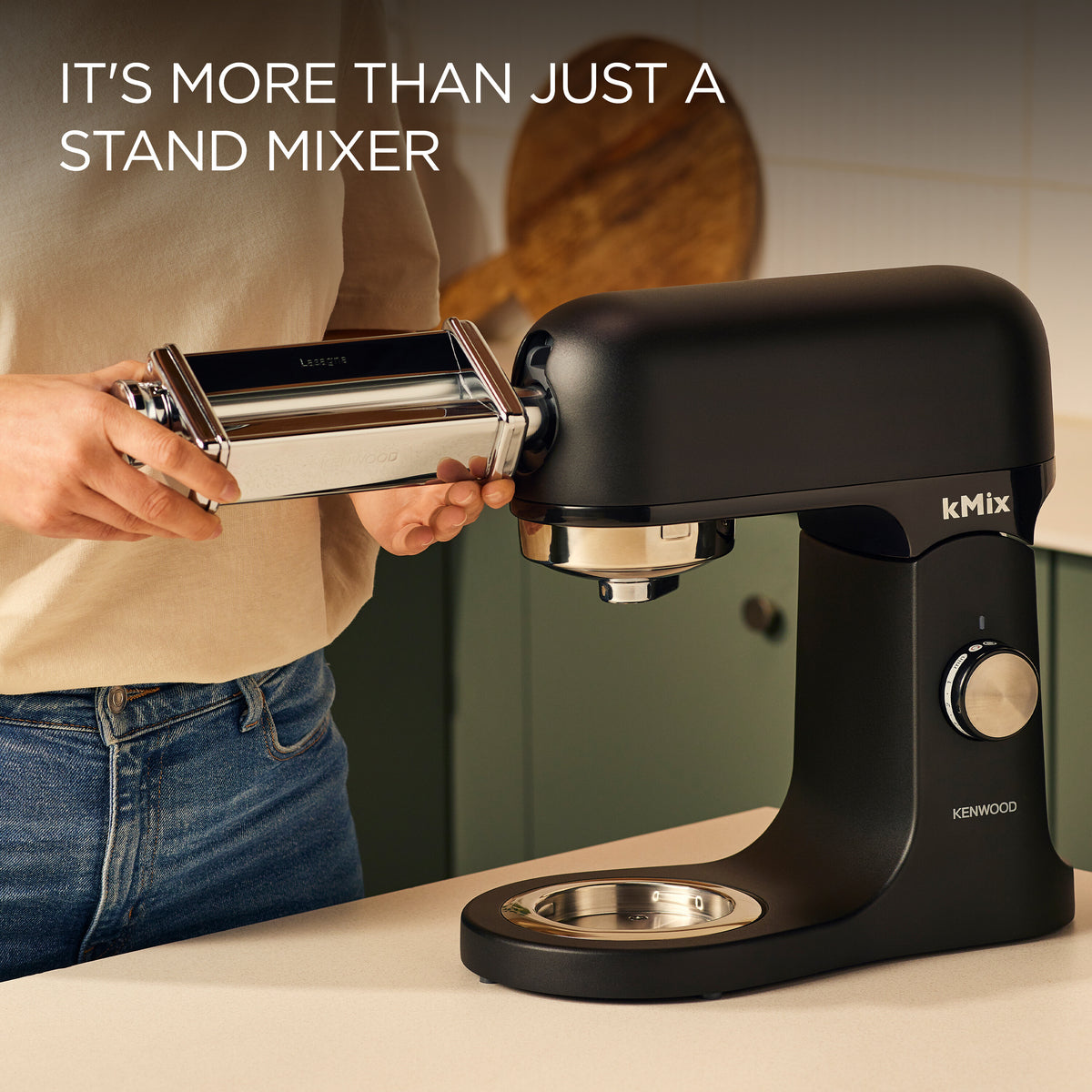 Close-up of a person attaching a pasta roller accessory to a black Kenwood kMix stand mixer on a kitchen counter.