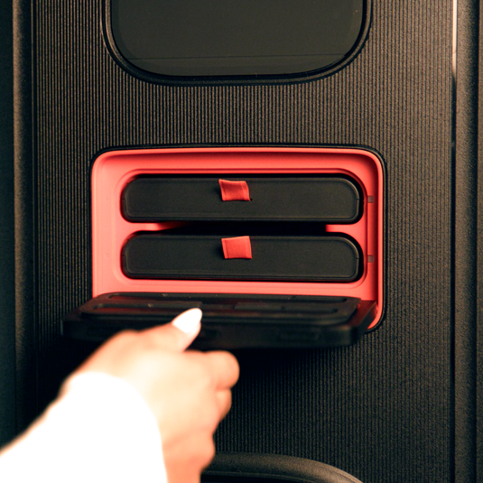 Hand holding open a black case revealing two JBL Battery 600 Duo batteries inside a red compartment.