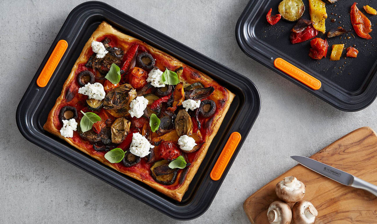Le Creuset 32cm rectangular baking sheet with vegetable pizza and roasted vegetables on a gray surface.