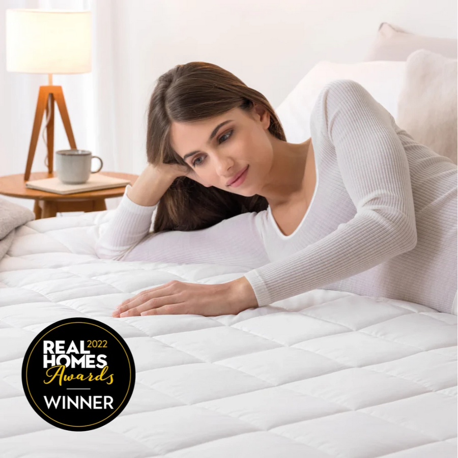 Woman lying on a white quilted Dreamland Snowed In Organic Cotton Warming Mattress Protector King with bedside lamp and cup