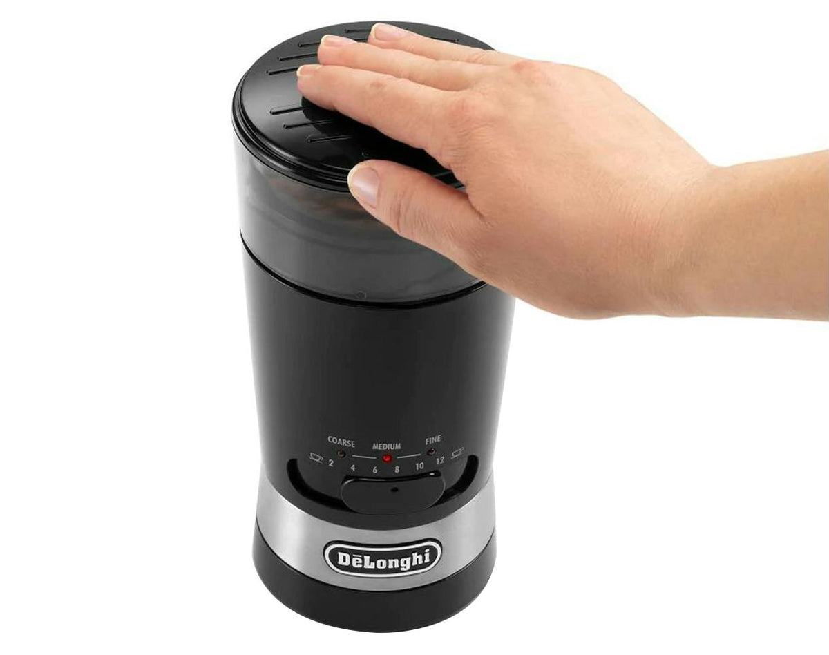 Hand pressing the top of a black DeLonghi Electric Blade Coffee Grinder KG210 with grind settings visible
