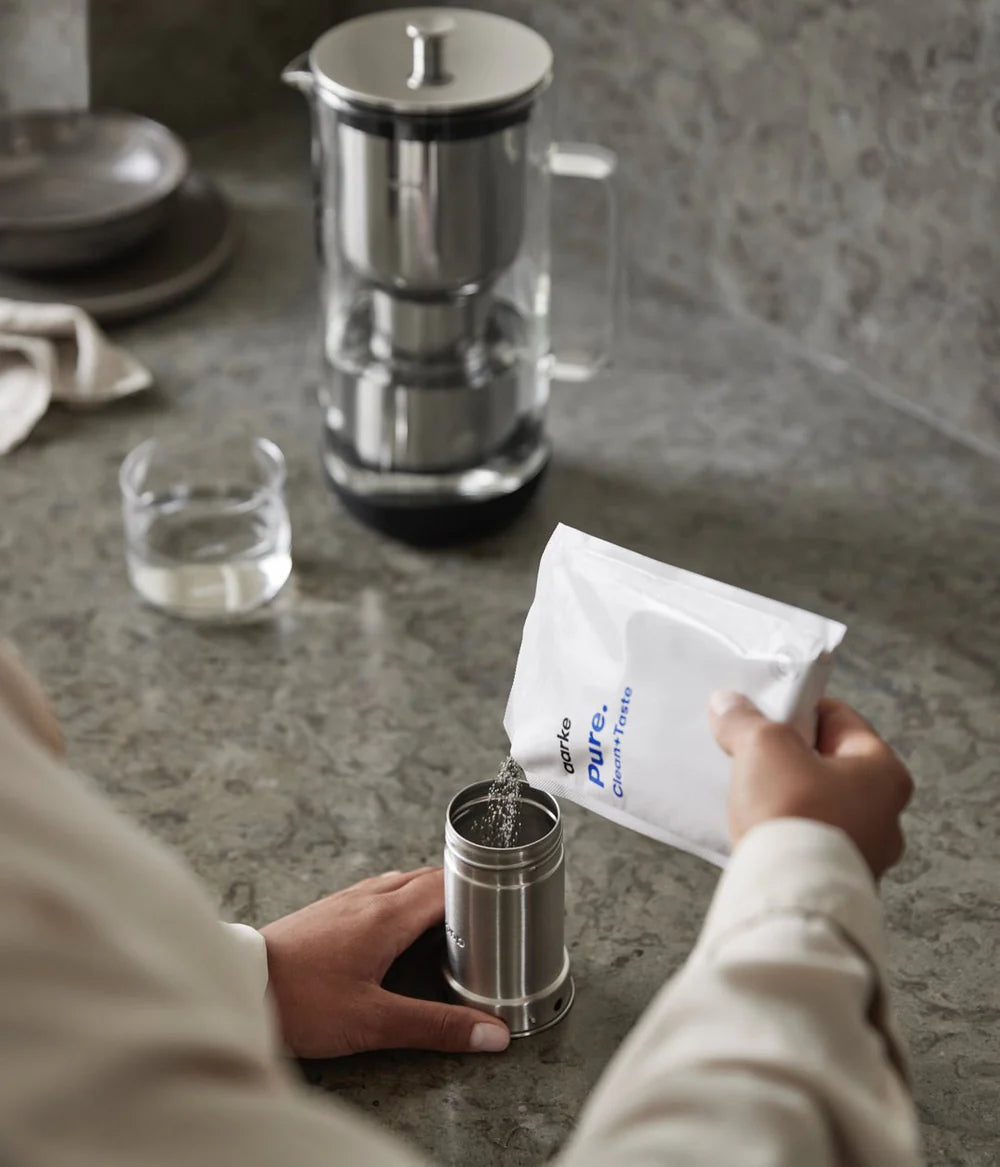 Person pouring Aarke Pure filter granules into a stainless steel filter container on a kitchen counter.