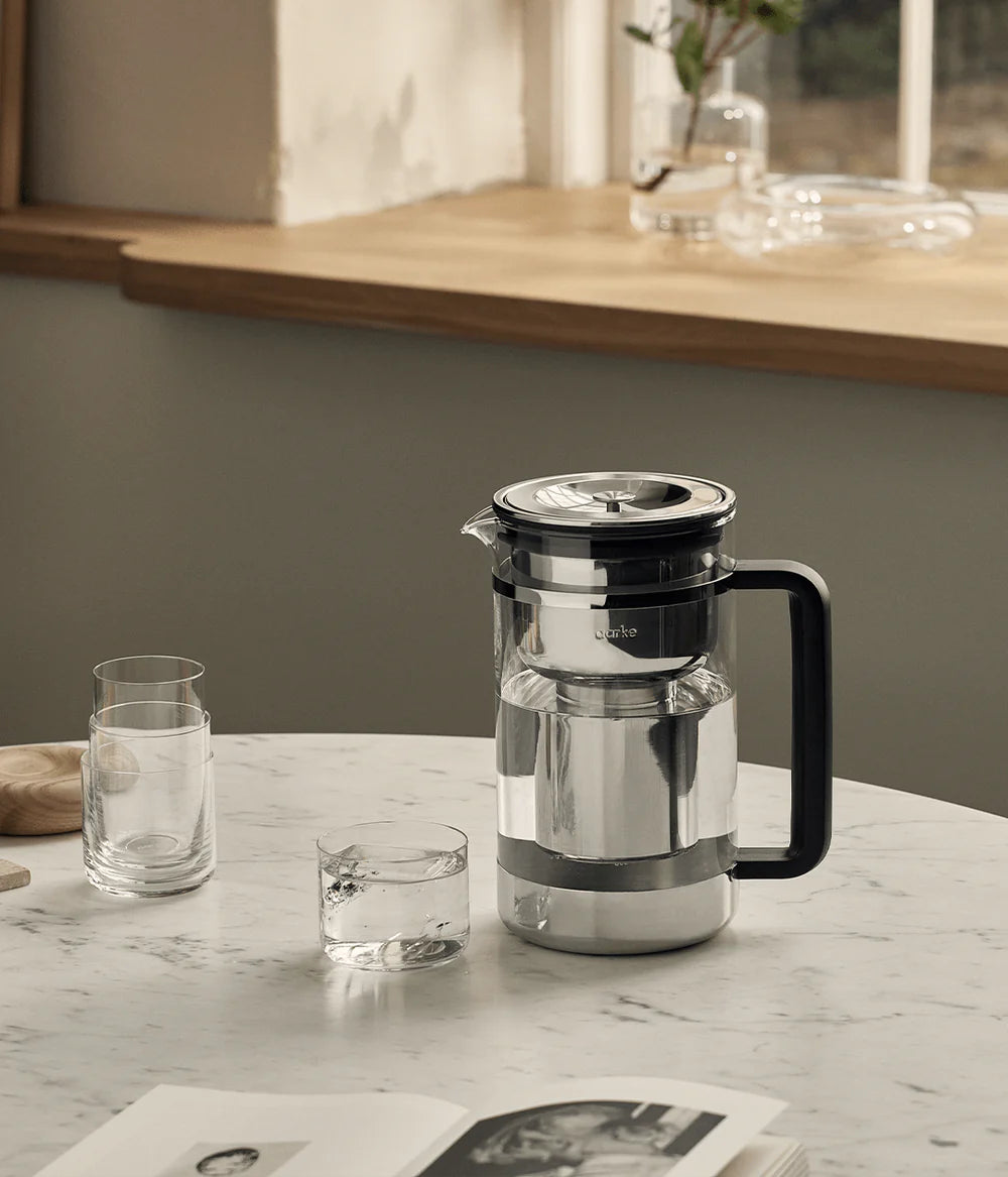 Aarke Purifier large glass water filter pitcher with black handle on a marble table next to glasses and an open book