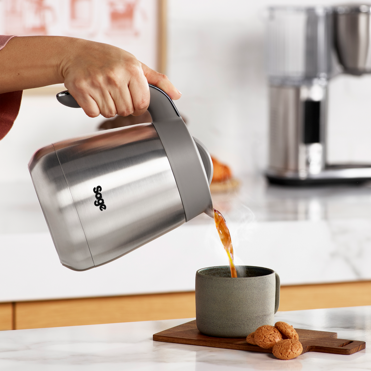 Hand pouring coffee from Sage Precision Brewer Thermal Brushed Steel into a gray mug on a kitchen counter.