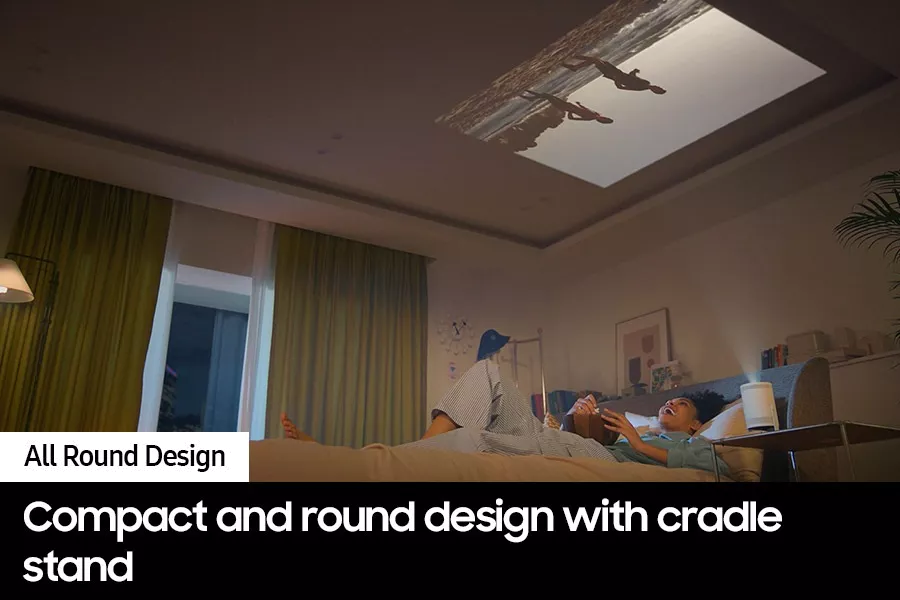 Samsung The Freestyle 2nd Gen projector on a side table projecting a beach scene onto the ceiling above a person lying on a