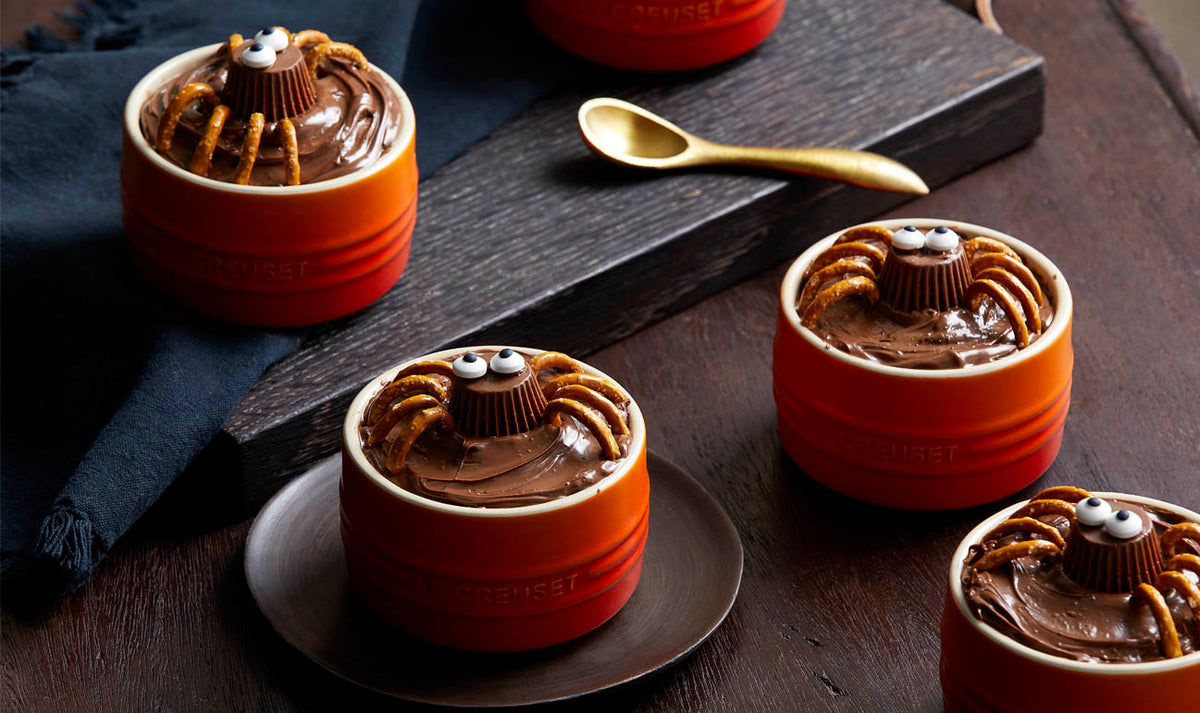 Four orange Le Creuset Ramekins filled with chocolate dessert decorated as spiders with pretzel legs and candy eyes.