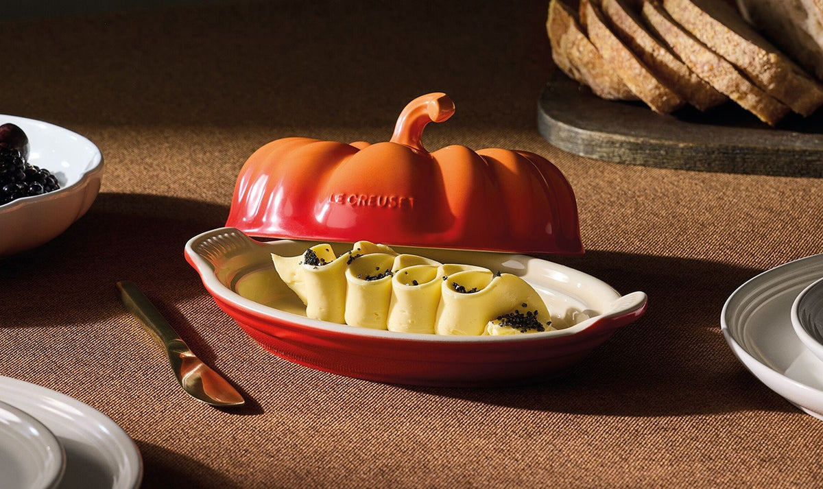 Le Creuset Halloween Pumpkin Butterdish in volcanic orange with butter curls inside on a brown tablecloth.