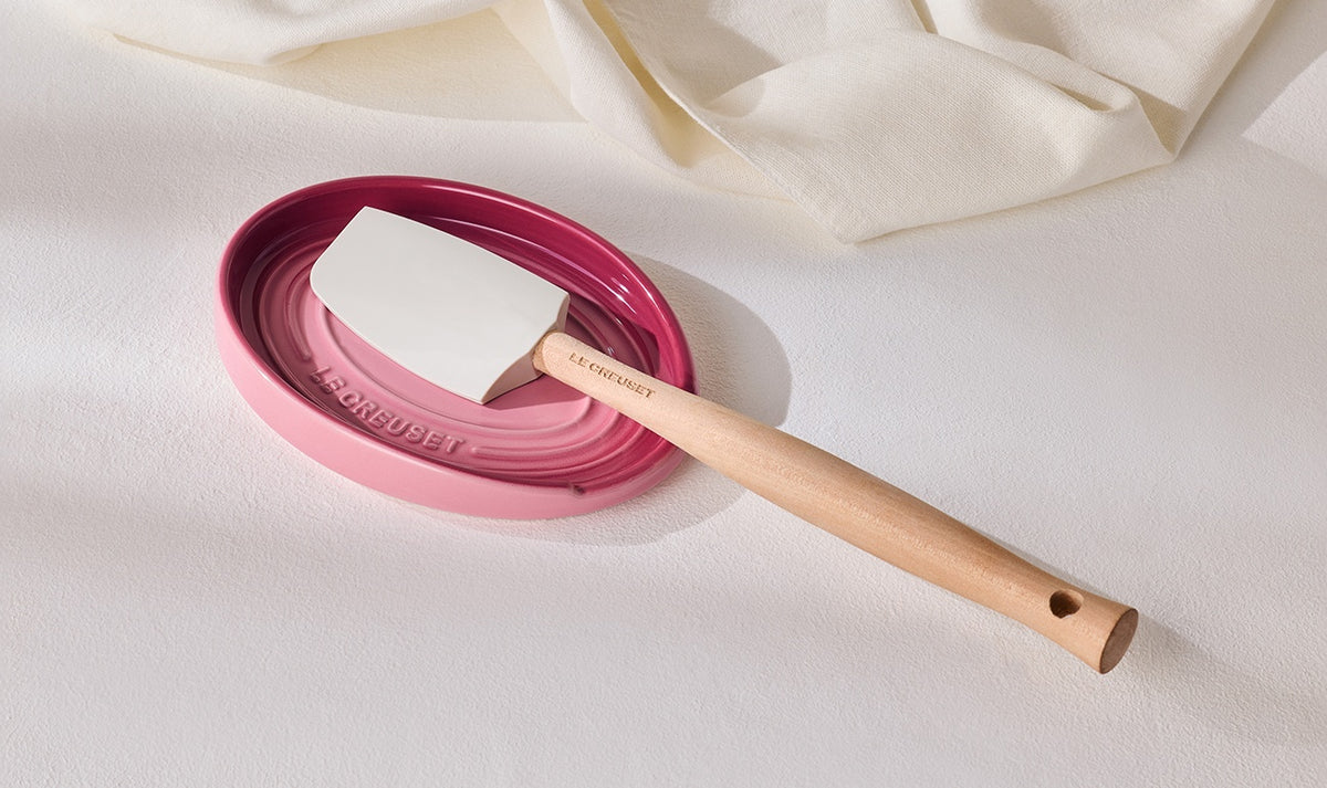 Le Creuset oval rose quartz spoon rest with a wooden-handled spatula on a white surface