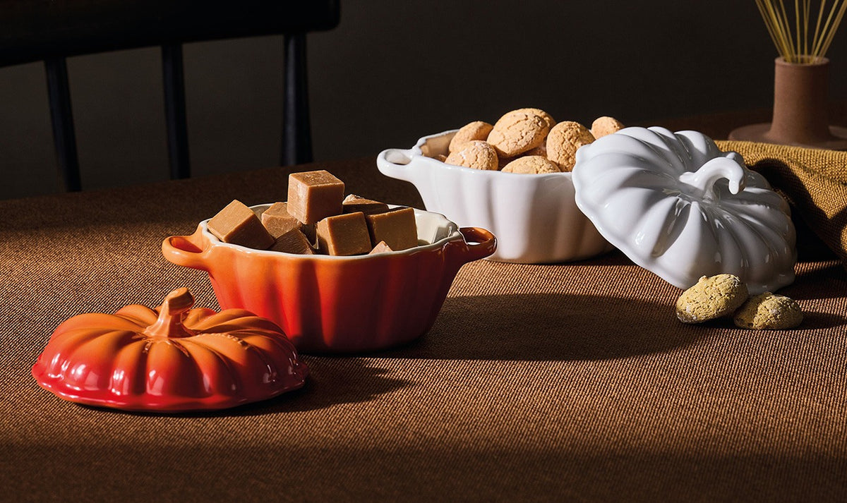 Two Le Creuset Halloween Pumpkin casseroles, one volcanic orange with caramel cubes and one white with cookies, both with