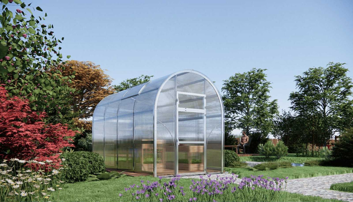 Greenhouse with arched roof and clear polycarbonate panels in a garden setting with flowers and trees.