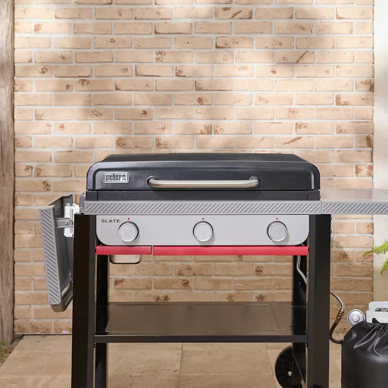Weber Slate GP 71cm Premium Griddle with three control knobs, side shelves, and propane tank against a brick wall.