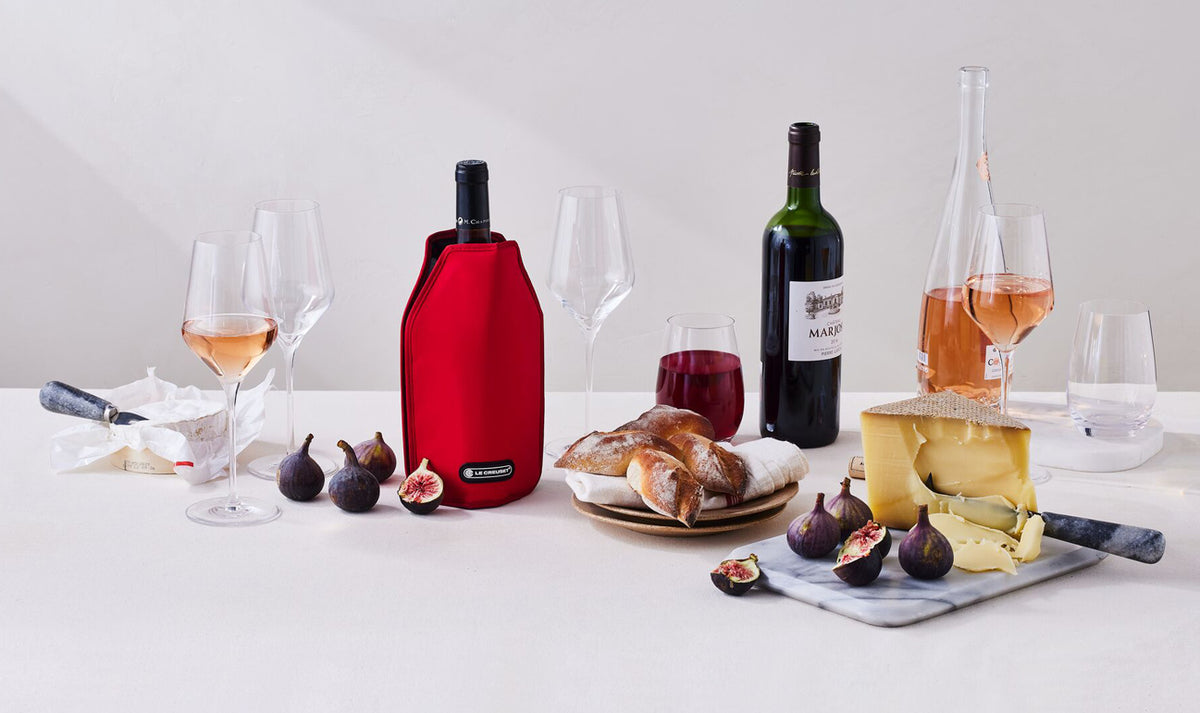 Le Creuset Cerise wine cooler sleeve on a bottle surrounded by wine glasses, figs, bread, and cheese on a table.