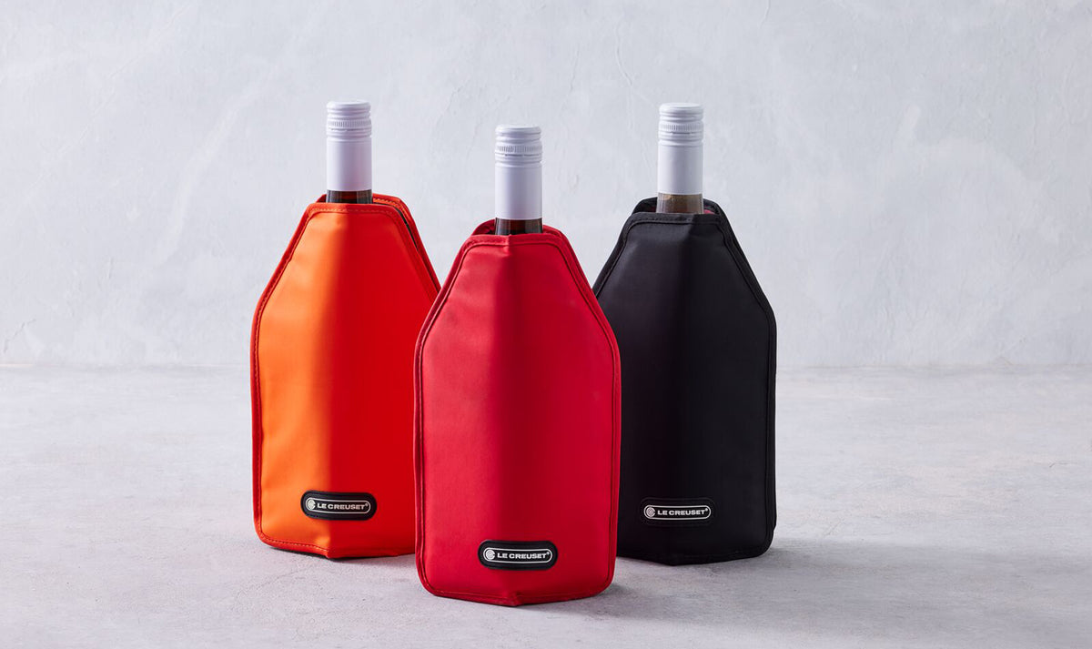 Three Le Creuset wine cooler sleeves in cerise red, orange, and black, each on a wine bottle.