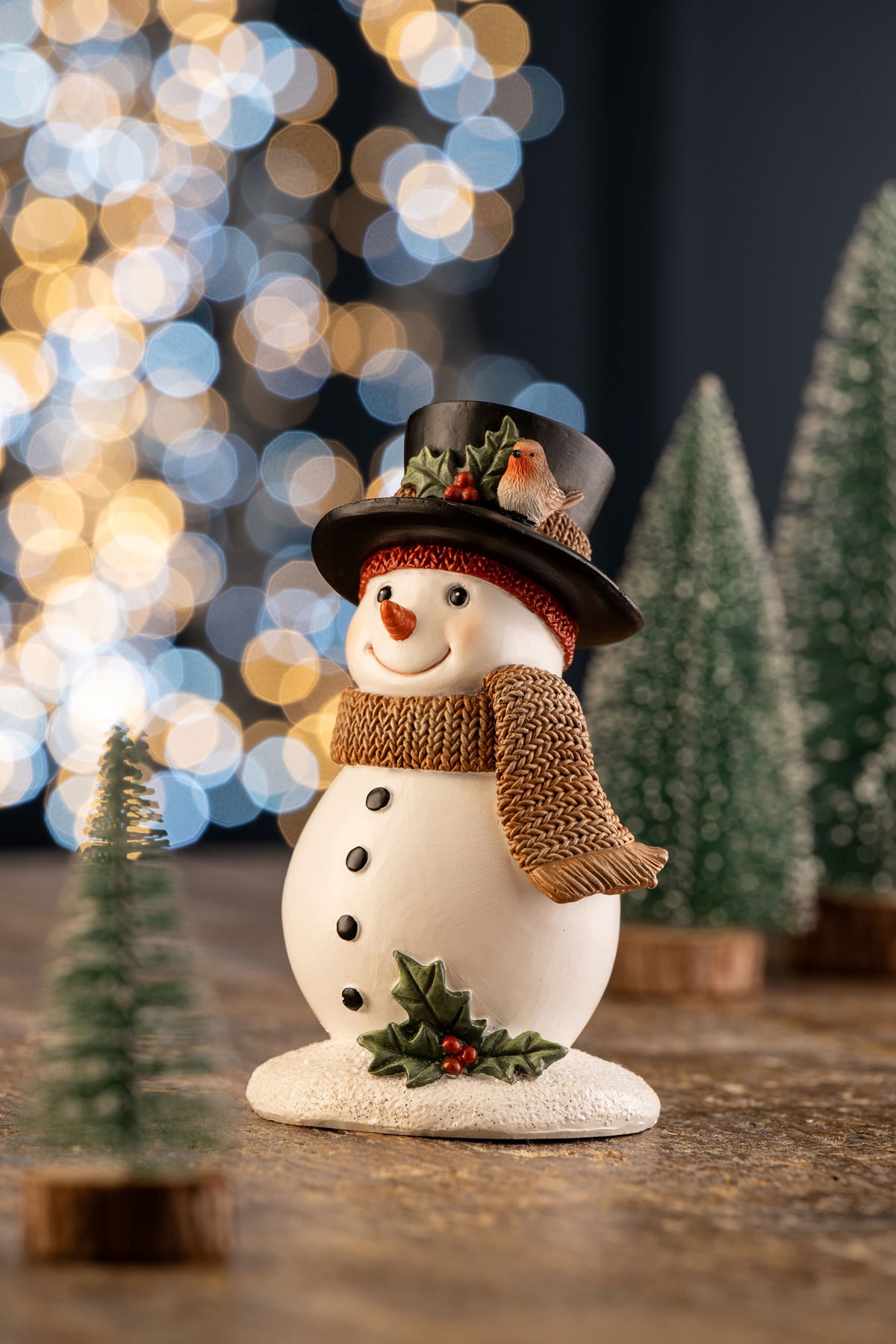 Snowman figurine wearing a black top hat with holly and a robin, and a brown knitted scarf, on a snowy base.