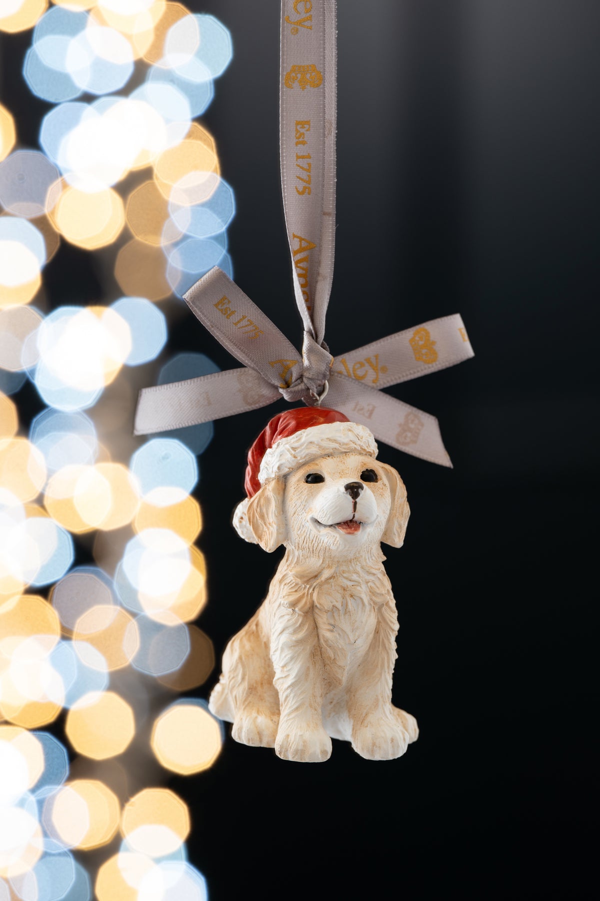 Aynsley Christmas Puppy ornament with a smiling dog wearing a red Santa hat, hanging by a branded ribbon.
