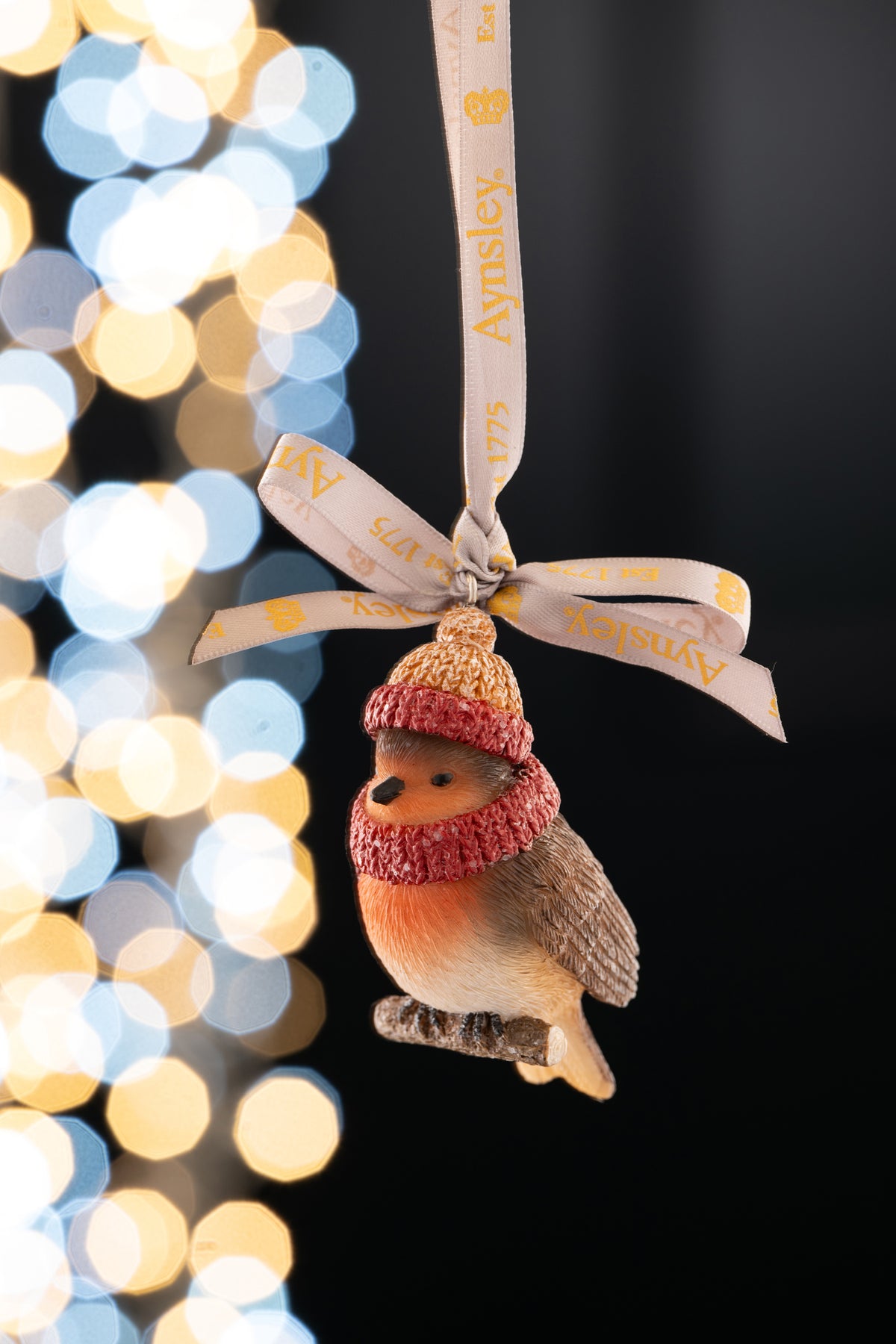 Aynsley robin ornament wearing a knitted woolly hat and scarf, hanging from a branded ribbon.