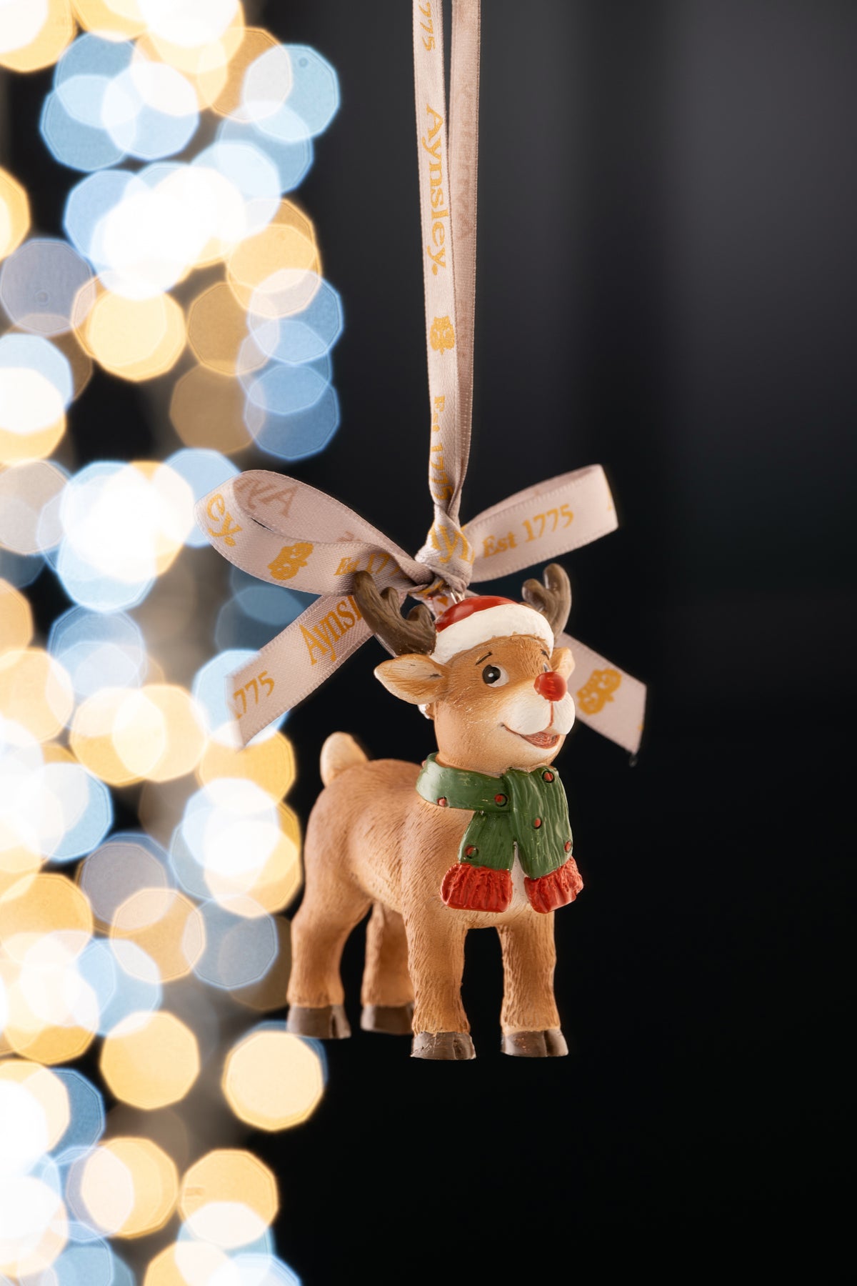 Aynsley Rudolph the Reindeer ornament wearing a Santa hat and green scarf, hanging by a branded ribbon.