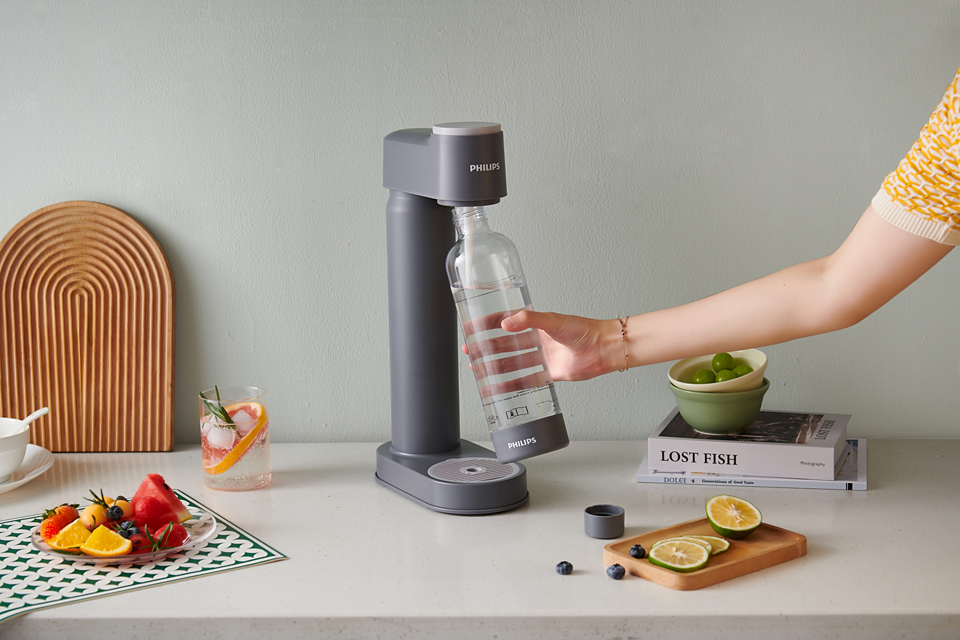 Philips GoZero Soda Maker in gray with a hand holding the attached clear bottle on a kitchen counter.