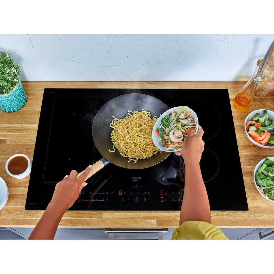 Beko 80cm induction hob in use with a person cooking noodles and vegetables in a wok on a wooden countertop.