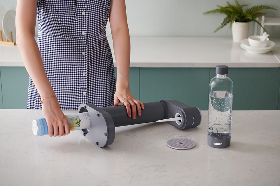 Philips GoZero Soda Maker in dark gray with a person inserting a bottle, next to a carbonated water bottle on a kitchen