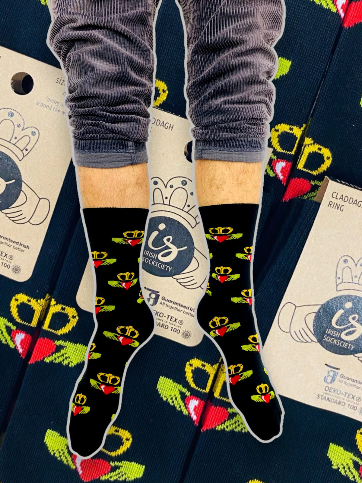 Legs wearing black Galway Claddagh Ring socks with red heart and yellow crown pattern on packaging background.