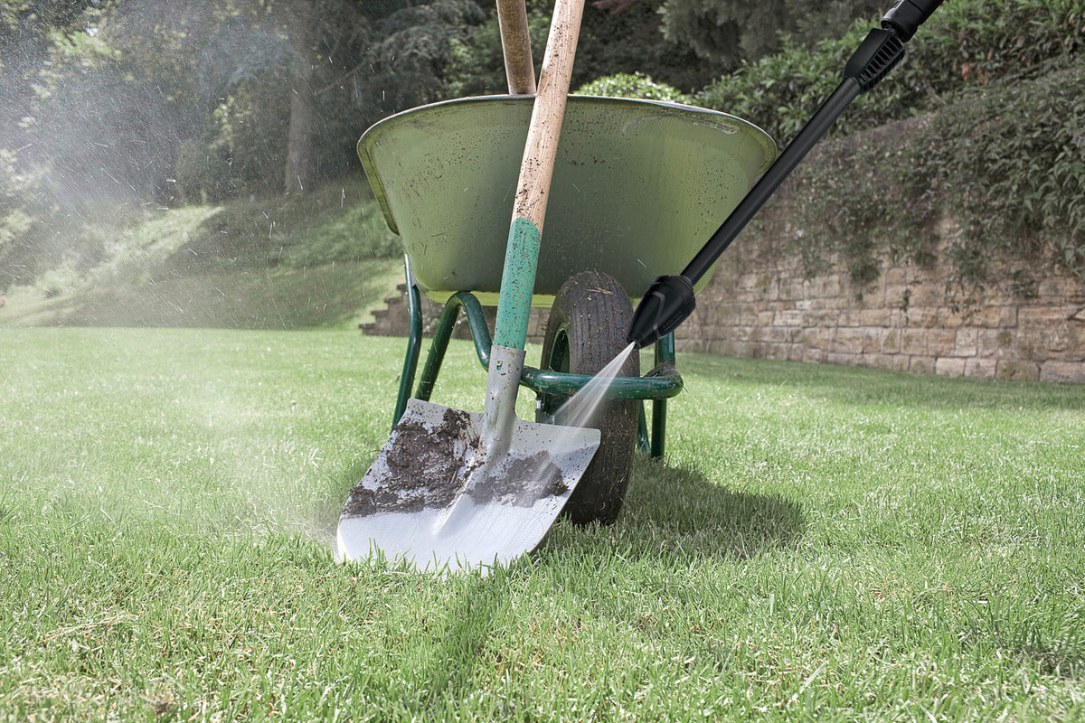 Karcher Full Control Dirt Blaster nozzle spraying water on a muddy shovel leaning against a wheelbarrow on grass.