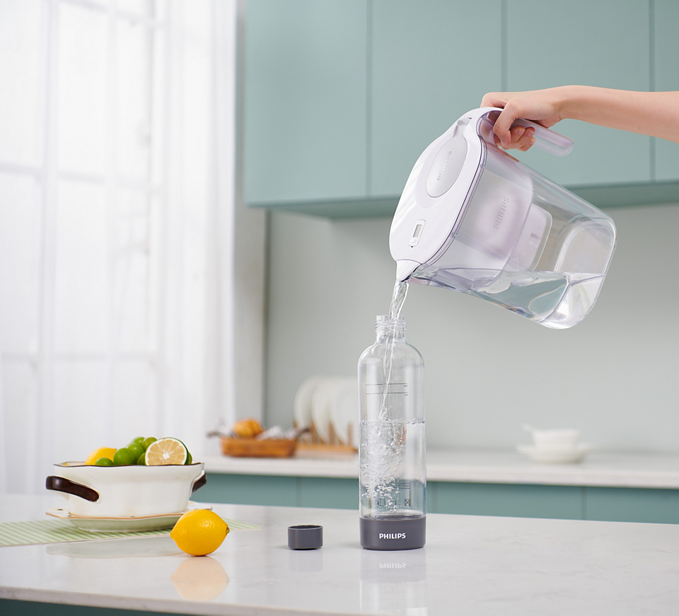 Hand pouring water from Philips GoZero Soda Maker pitcher into a clear bottle on kitchen counter.
