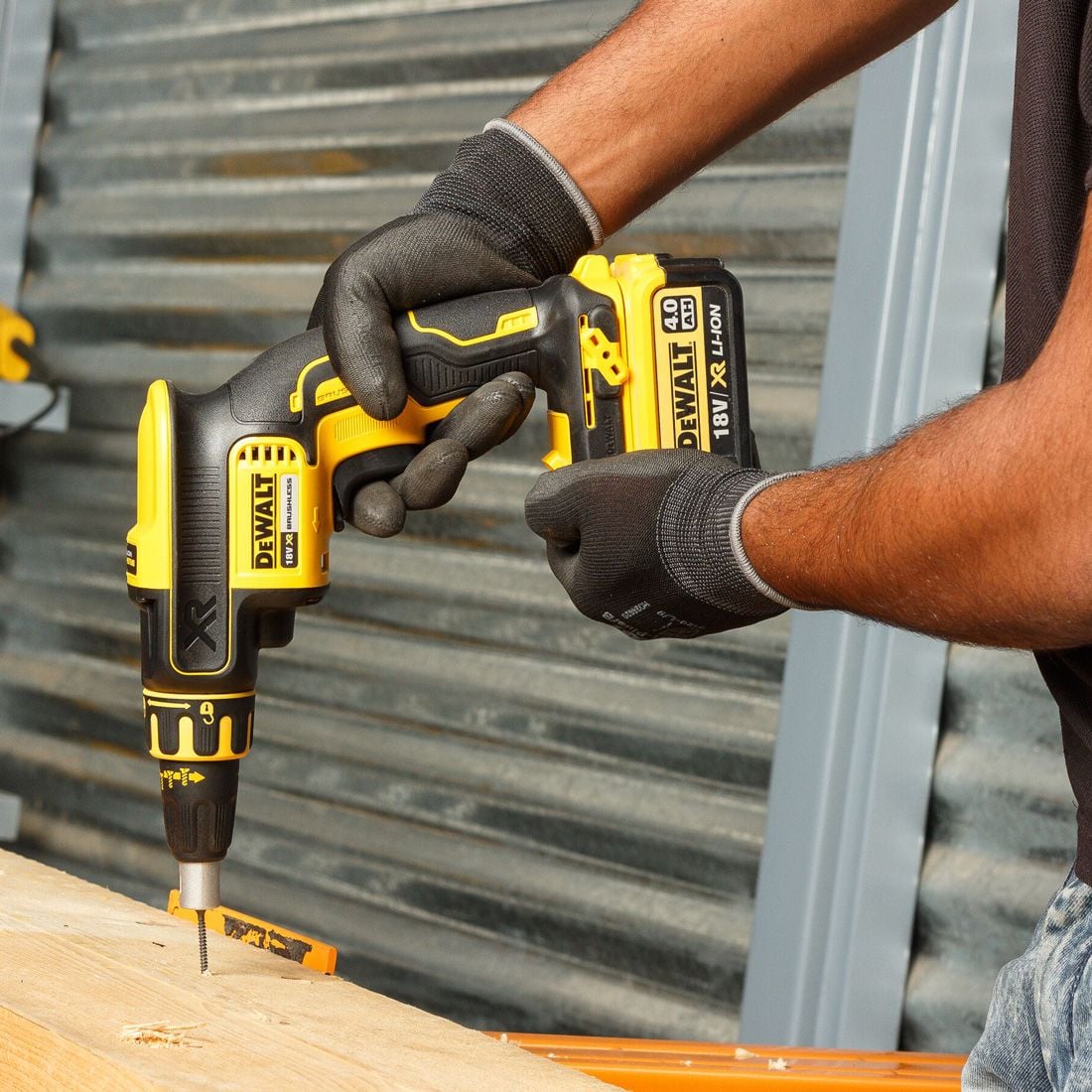 A person wearing black work gloves uses a yellow and black DeWalt cordless drill to drive a screw into a piece of wood, with a corrugated metal wall in the background.