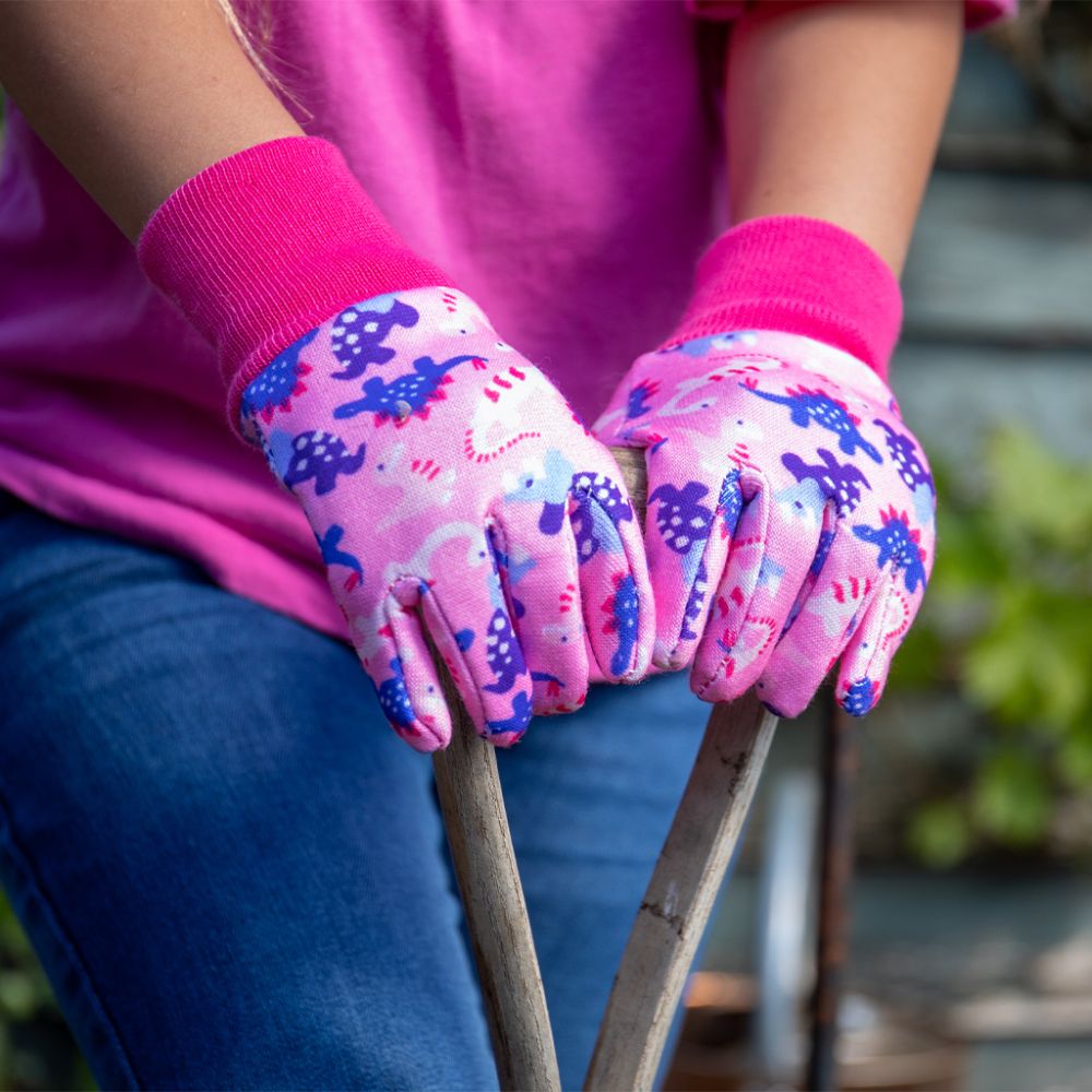 Child wearing pink Kent & Stowe dinosaur gardening gloves gripping wooden garden tool handles.