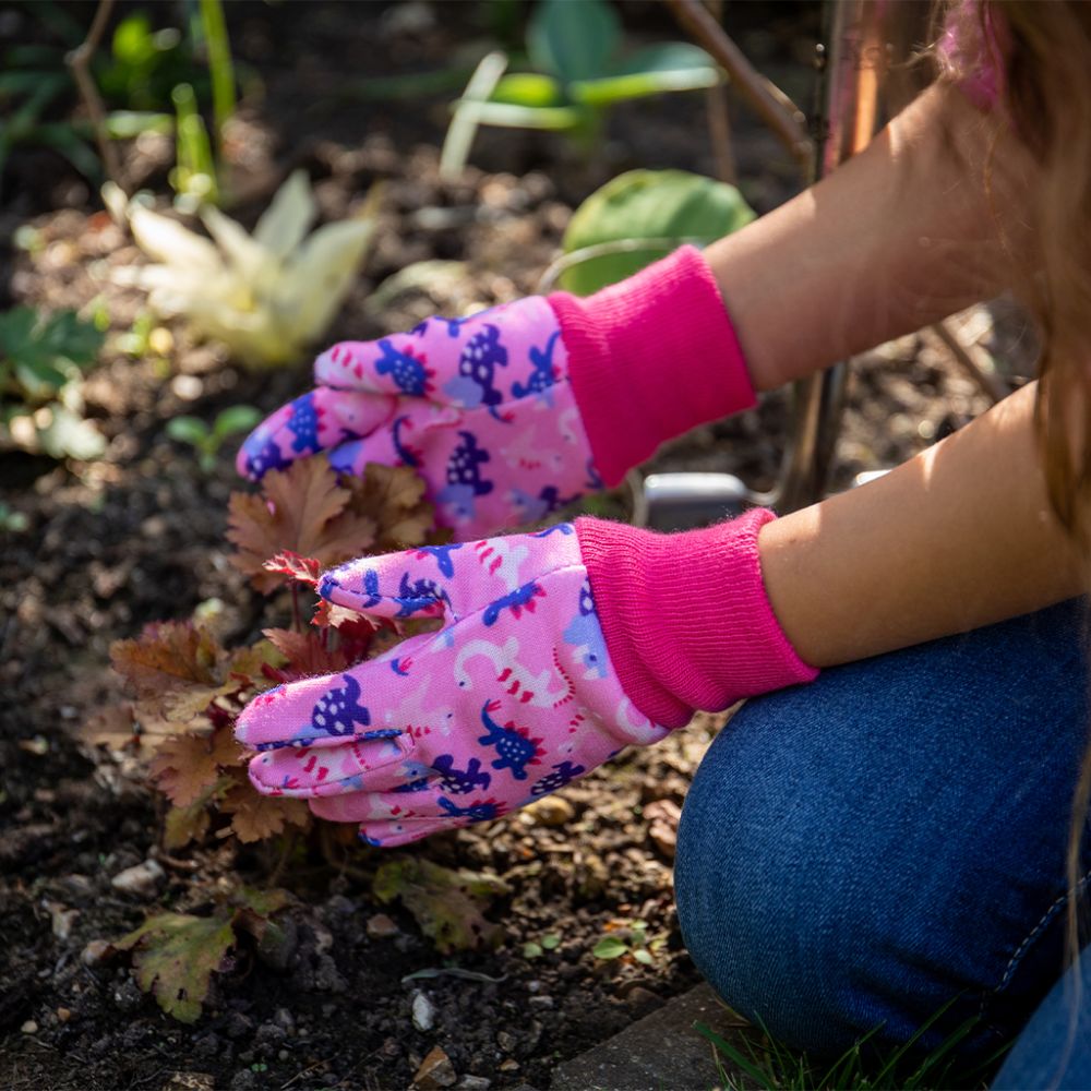 Child wearing pink Kent & Stowe dinosaur gardening gloves while tending plants in a garden bed.