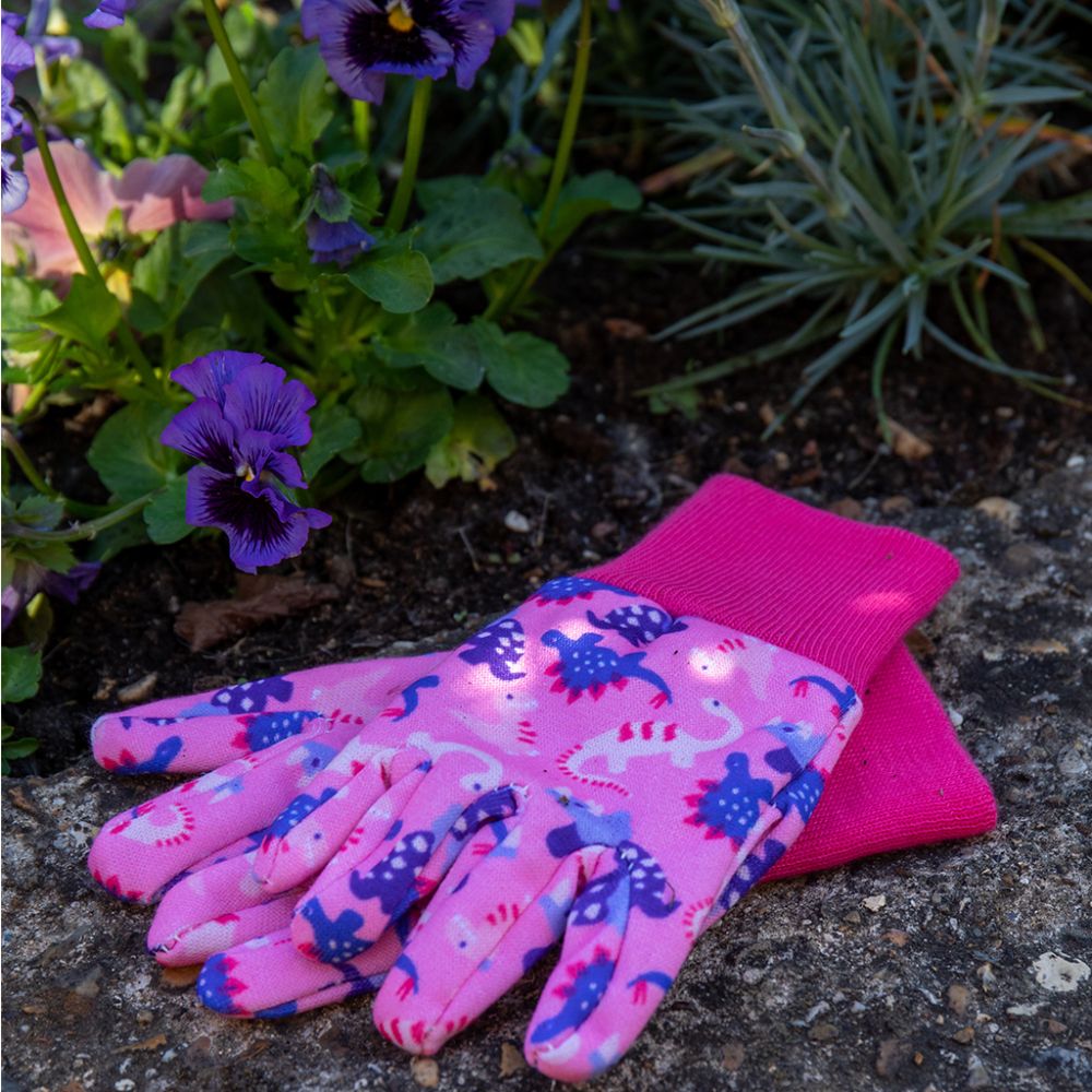 Pink kids gardening gloves with blue and white dinosaur patterns, shown outdoors near flowers and soil.