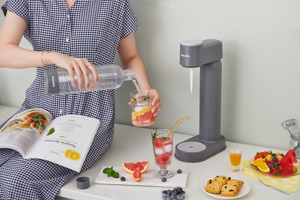 Philips GoZero Soda Maker in gray on a counter with a person pouring sparkling water into a glass with fruit and ice.