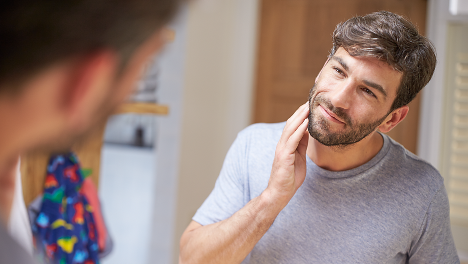 Man with trimmed beard touching his face in front of a mirror, suggesting grooming with Philips Multigroom series 3000 kit